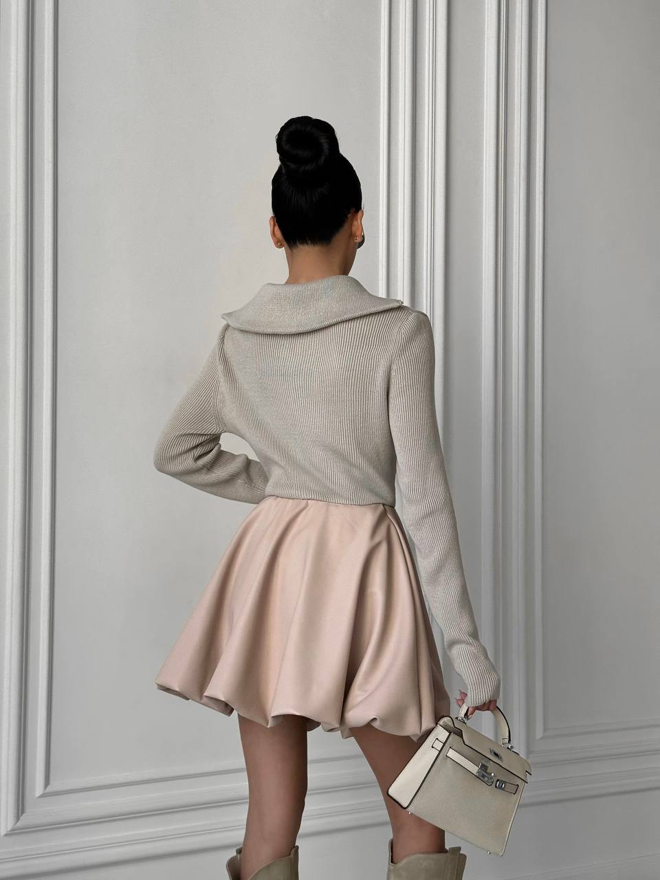 Woman in beige sweater and pink skirt holding a handbag against a gray wall.