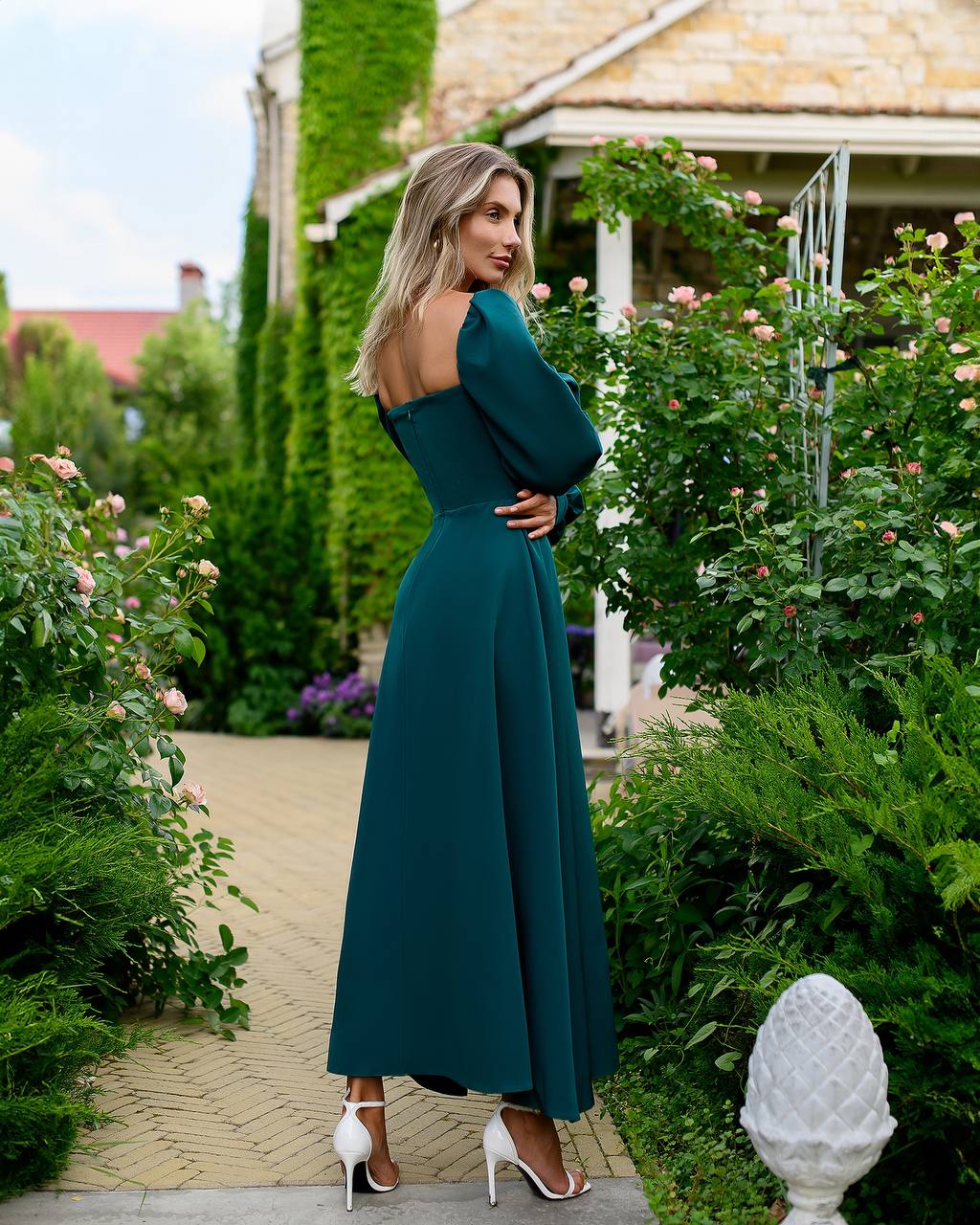 Woman in a teal dress standing in a garden with greenery and flowers around.