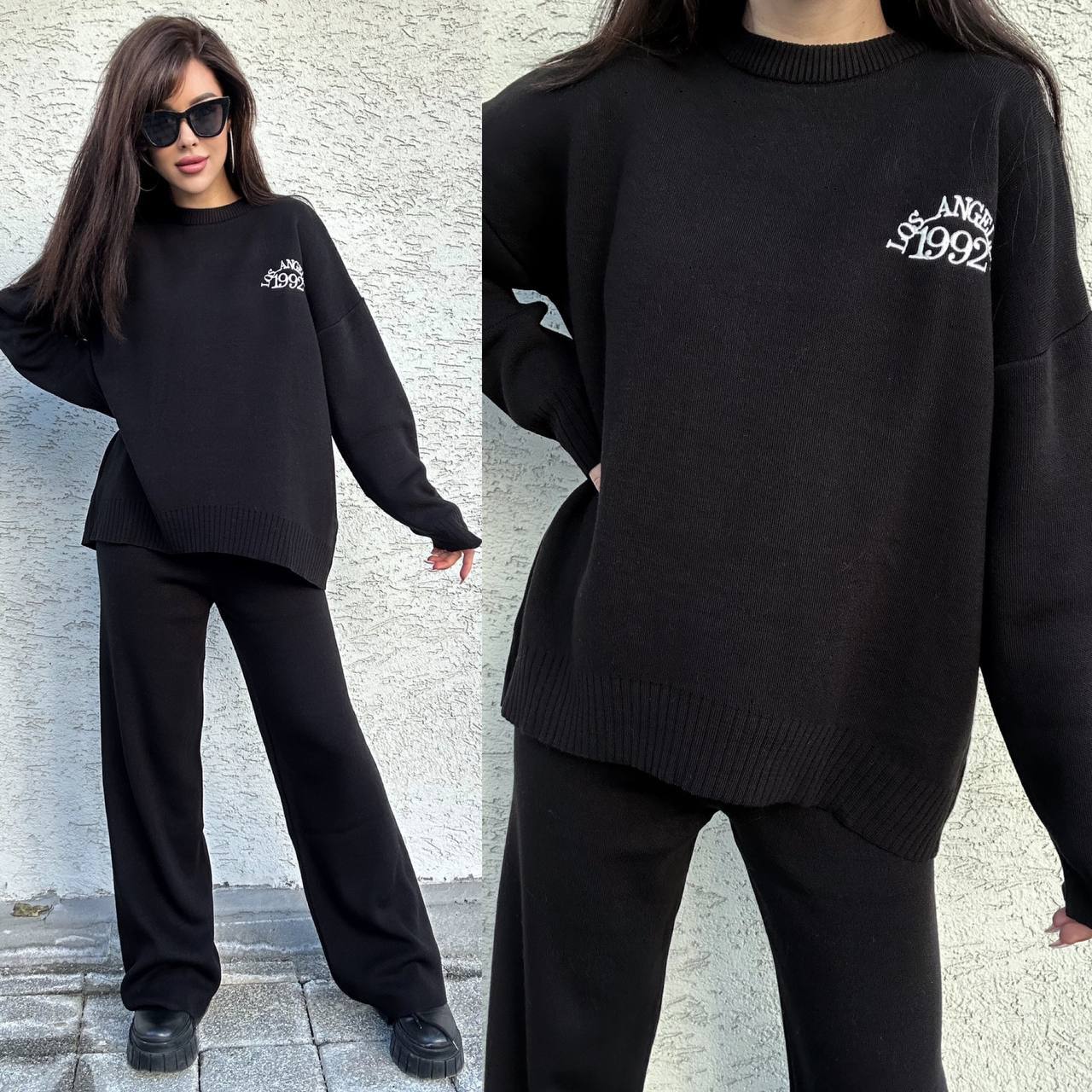 Woman wearing a black sweater and pants set with a brand logo, standing against a light-colored wall.