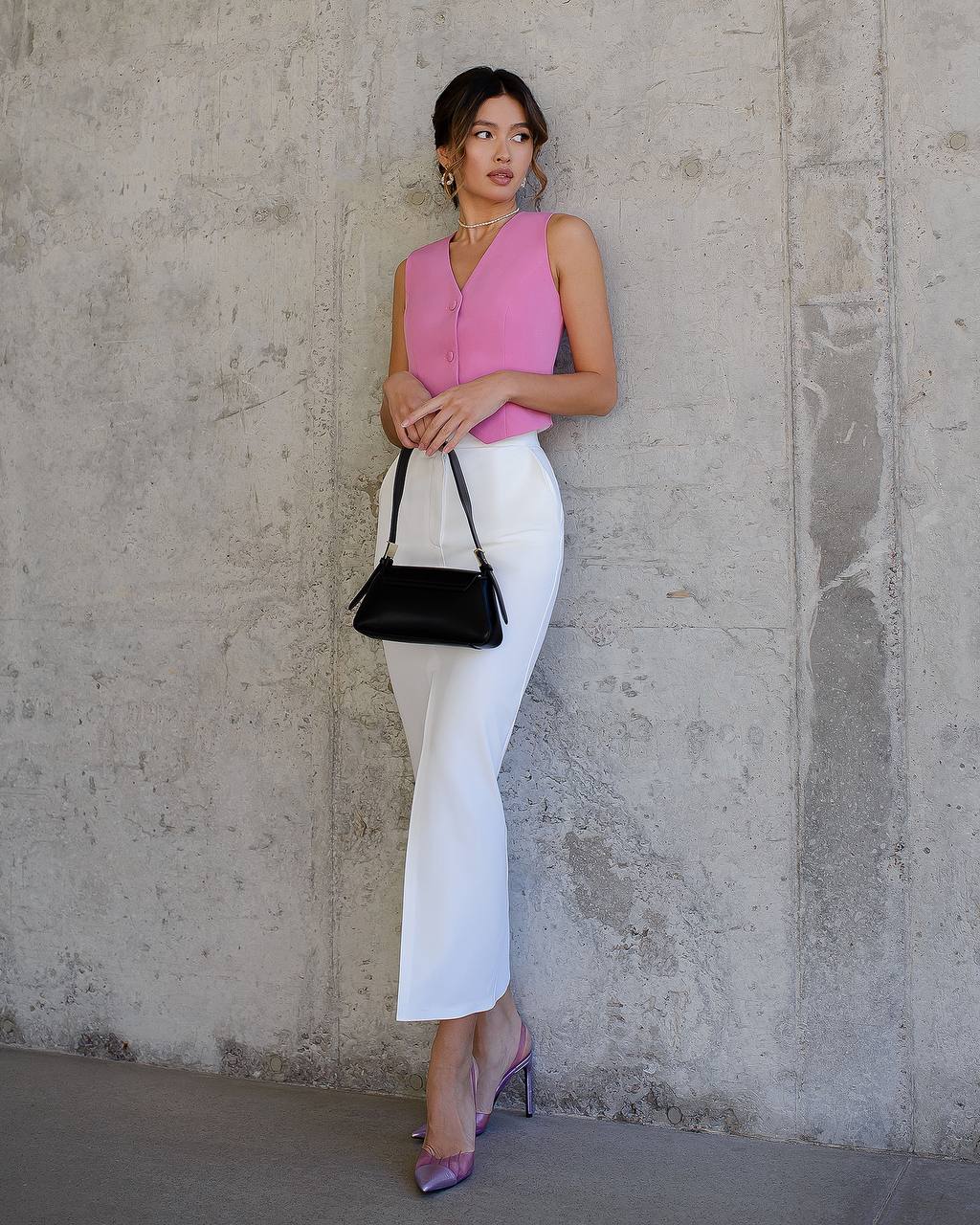 Woman in pink top and white pants holding a black handbag against a concrete wall.