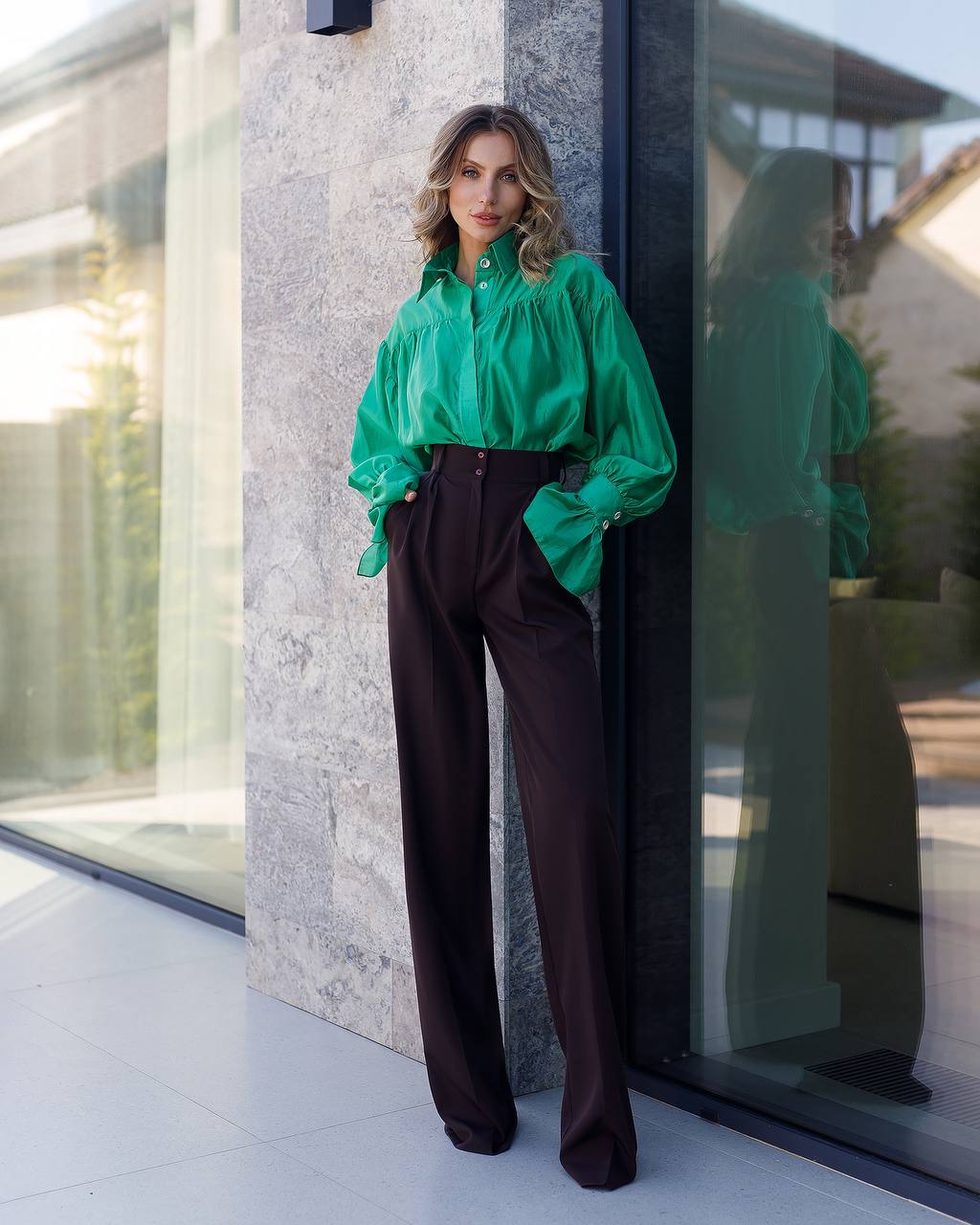 Woman in a green blouse and brown pants standing against a modern building.
