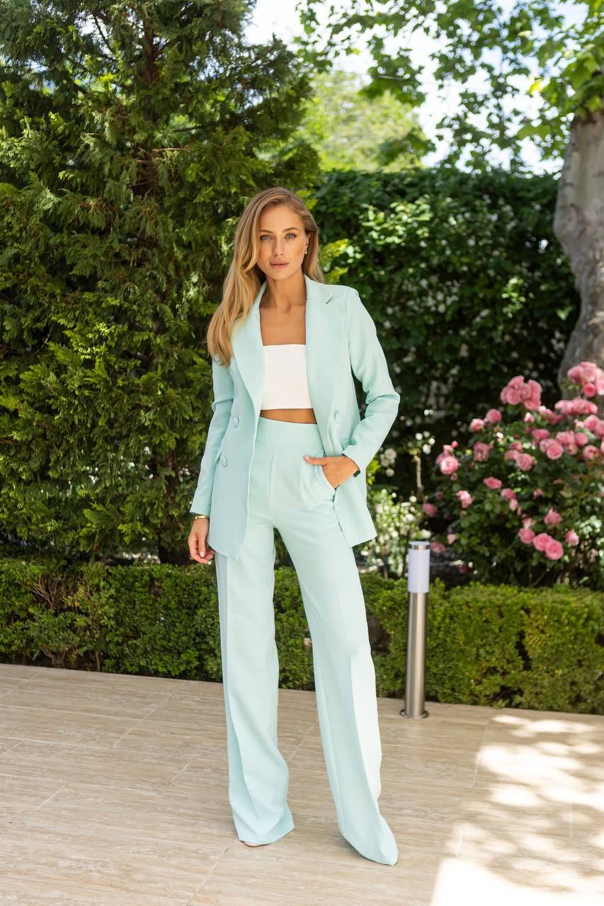 Woman in a light blue suit standing outdoors with greenery and flowers in the background