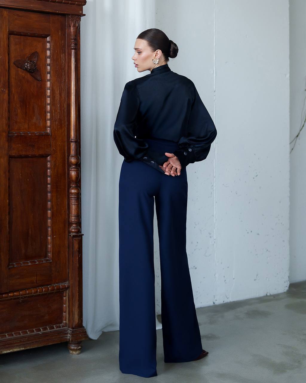 Woman in a navy outfit standing next to a wooden cabinet against a white wall.