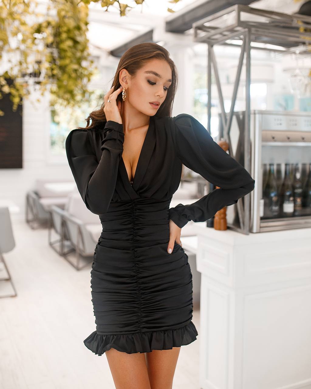 Woman in a black dress posing in an indoor setting with plants and furniture.