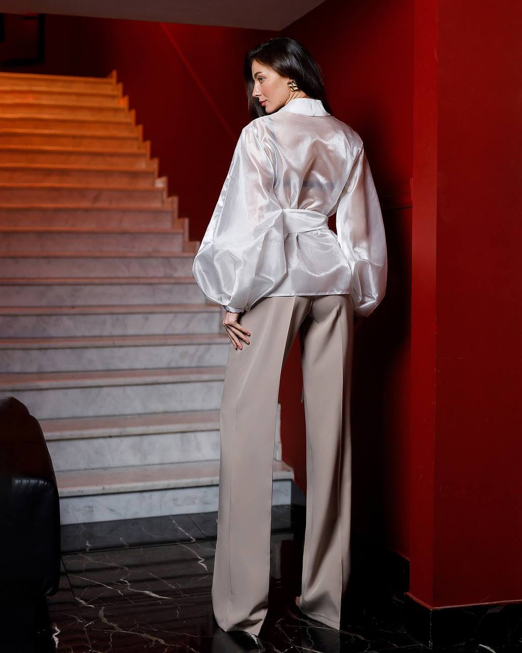Woman in a white satin outfit standing in a room with red walls and a staircase.