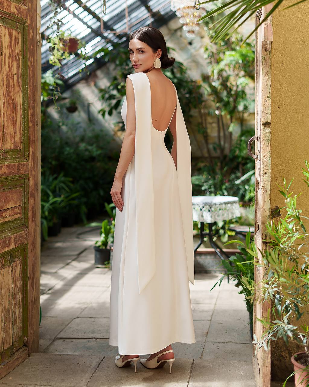 Woman in a white dress with a backless design standing in a greenhouse.