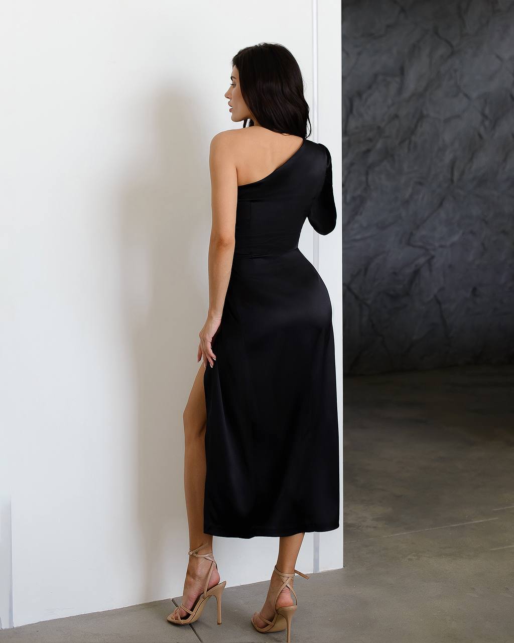 Woman wearing a black one-shoulder dress with a slit, standing against a white wall.