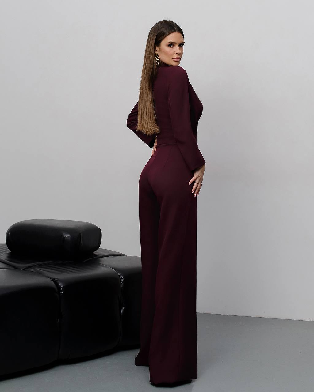 Woman in a burgundy jumpsuit standing in a minimalistic room with a black leather couch.