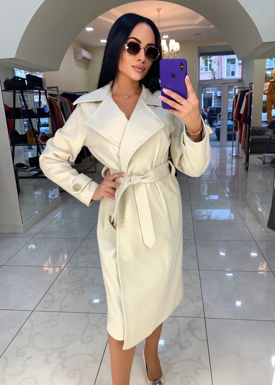 Woman taking a mirror selfie wearing a white trench coat in a store.