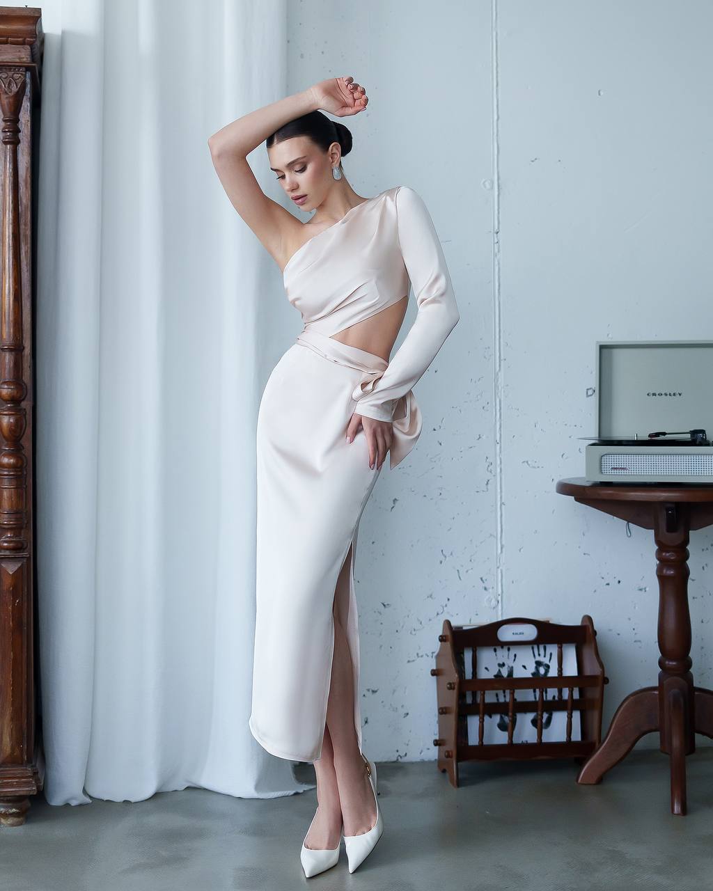 Woman in a white one-shoulder dress with a high slit, standing in a room with a record player and wooden furniture.