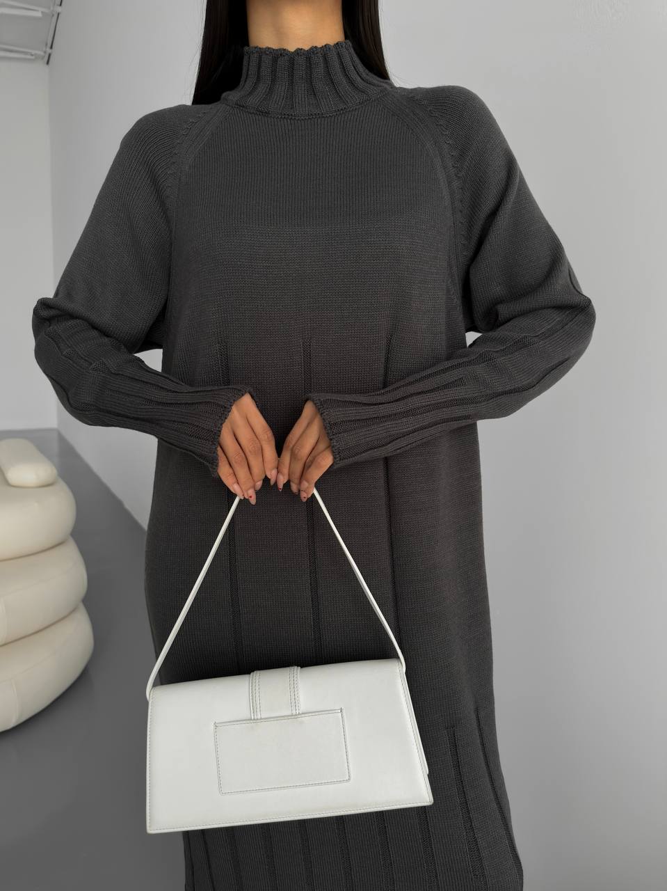 Person wearing a dark gray sweater holding a white handbag against a neutral background