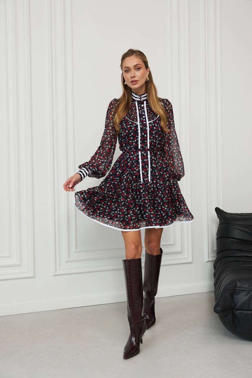 Woman wearing a floral dress with long sleeves and knee-high boots in a room with white paneled walls and a black leather chair.