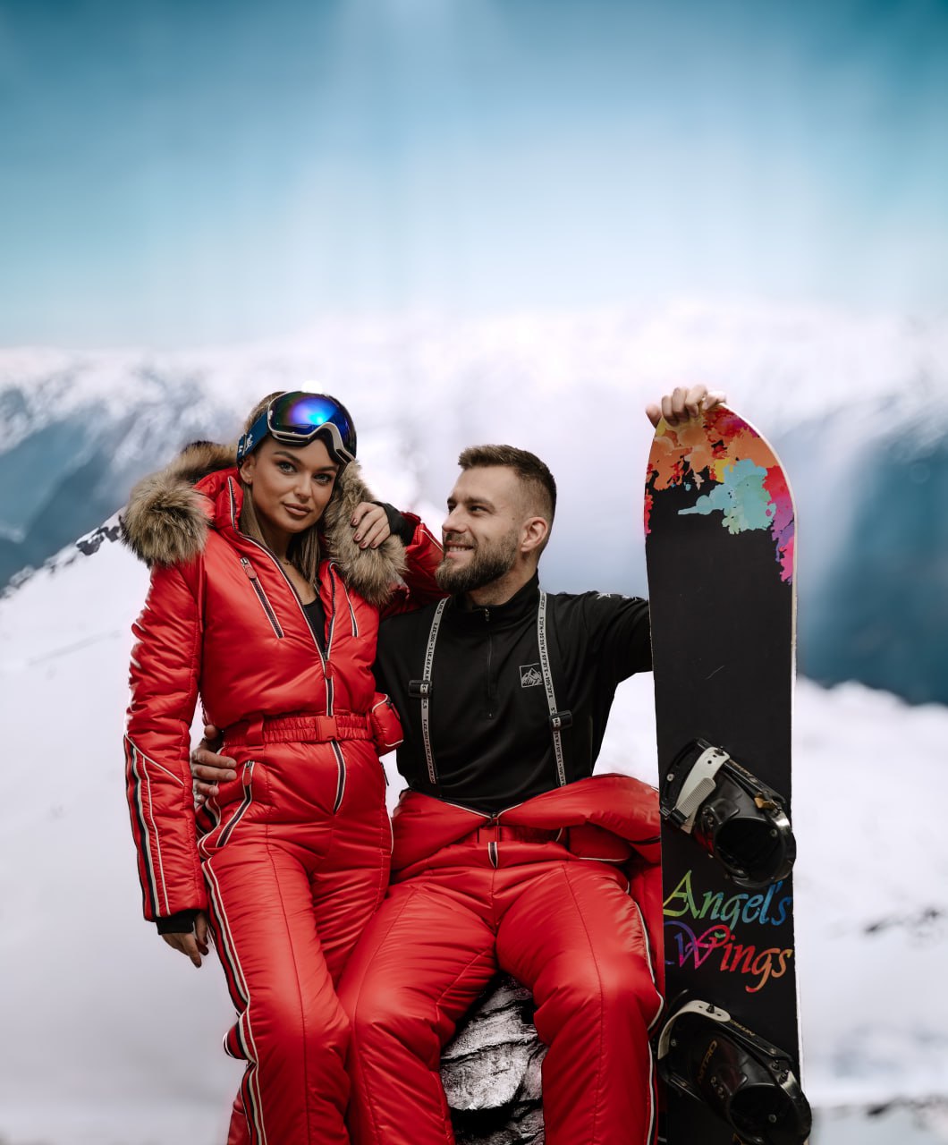 Two people in red ski suits with a colorful snowboard against a mountainous background