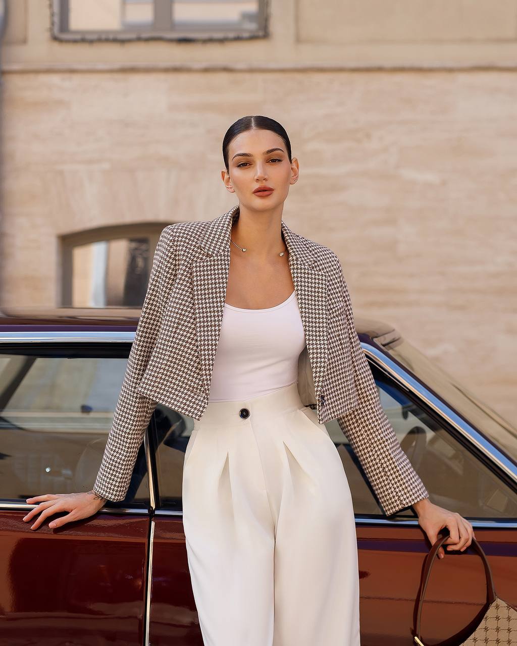 Woman in a checkered blazer and white pants standing next to a vintage car.