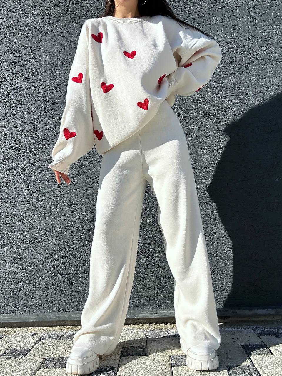 Person wearing a white sweater with red heart patterns and white pants against a gray wall.