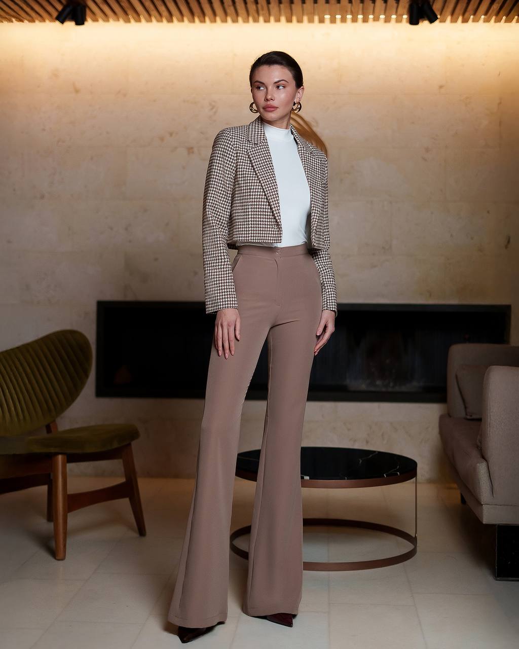 Woman in a checkered blazer and brown pants standing in a modern living room.