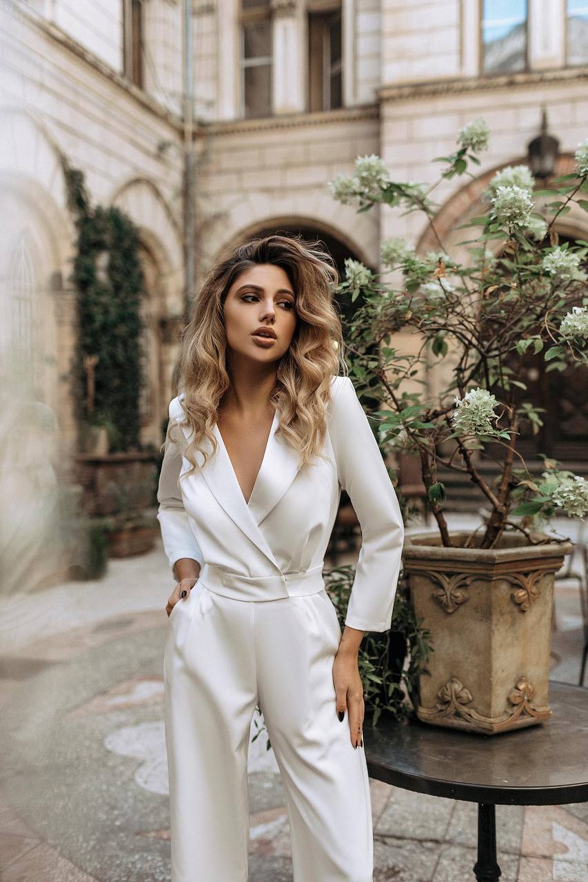 Woman in a white outfit standing in an outdoor setting with plants and architecture.