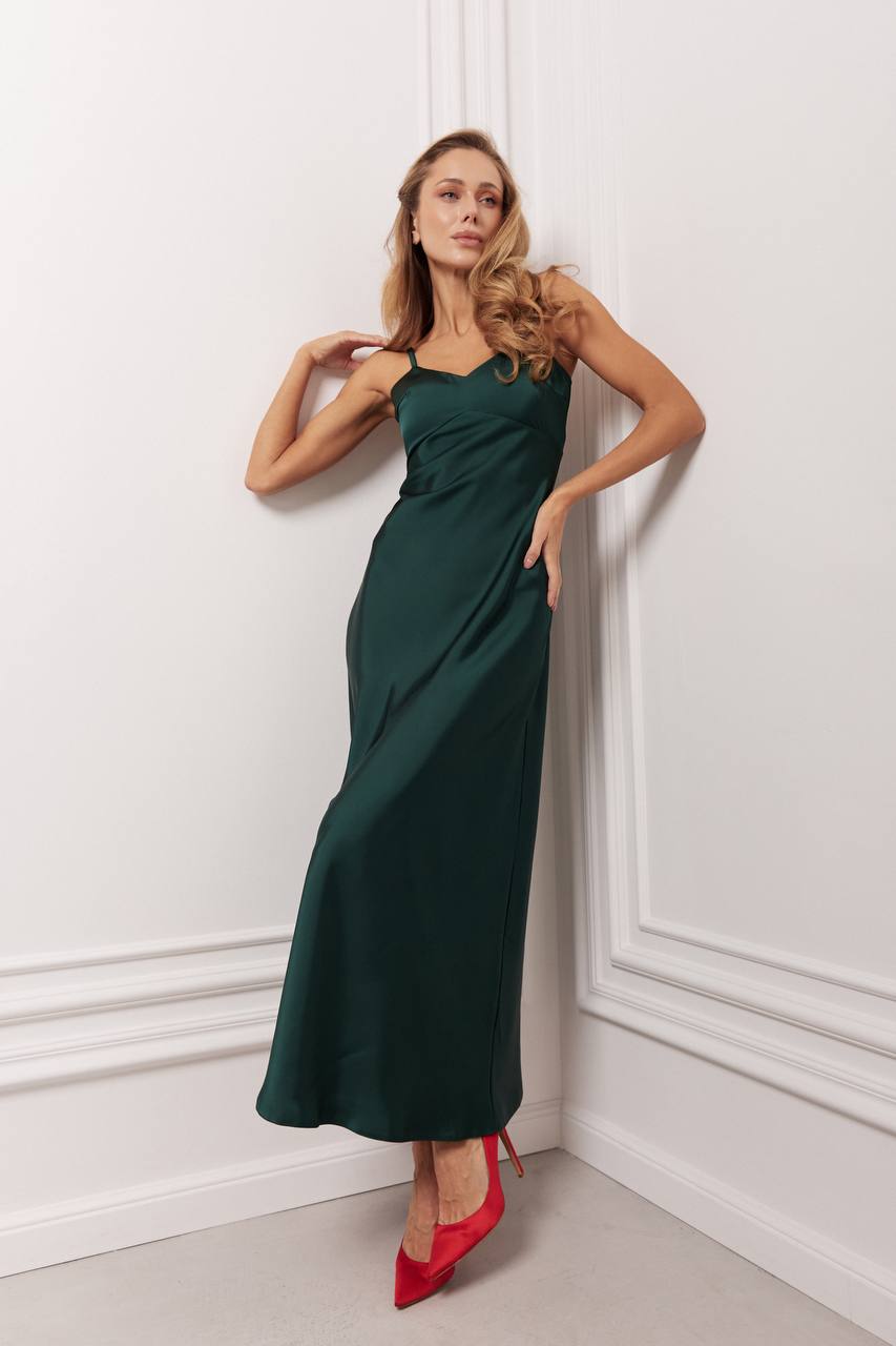 Woman in a dark green dress with red shoes standing against a white wall.