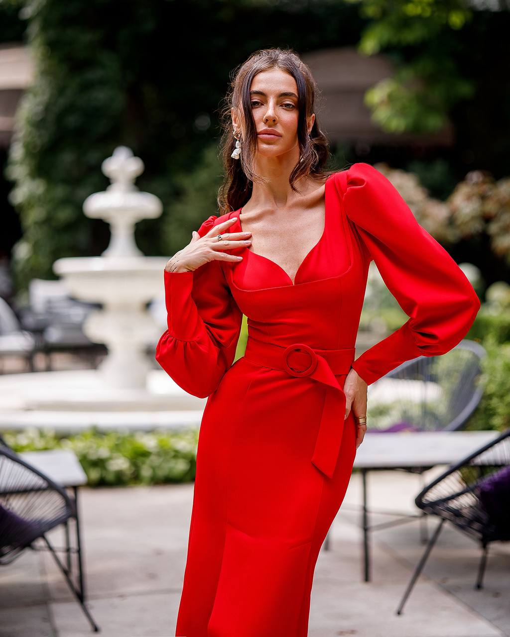 Woman in a red dress standing outdoors with greenery and a fountain in the background