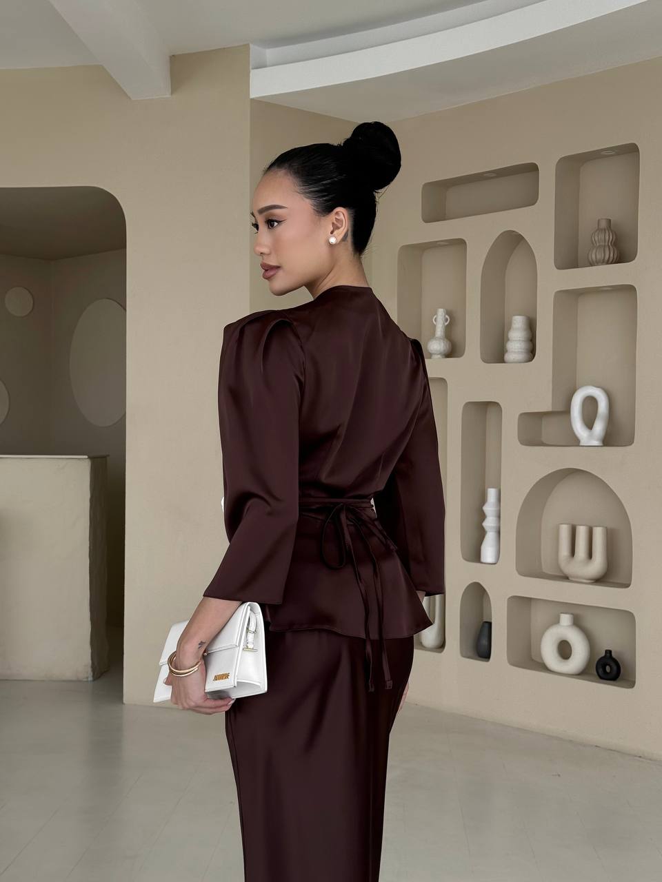 Woman in a brown outfit holding a white bag in a modern interior setting with shelves.
