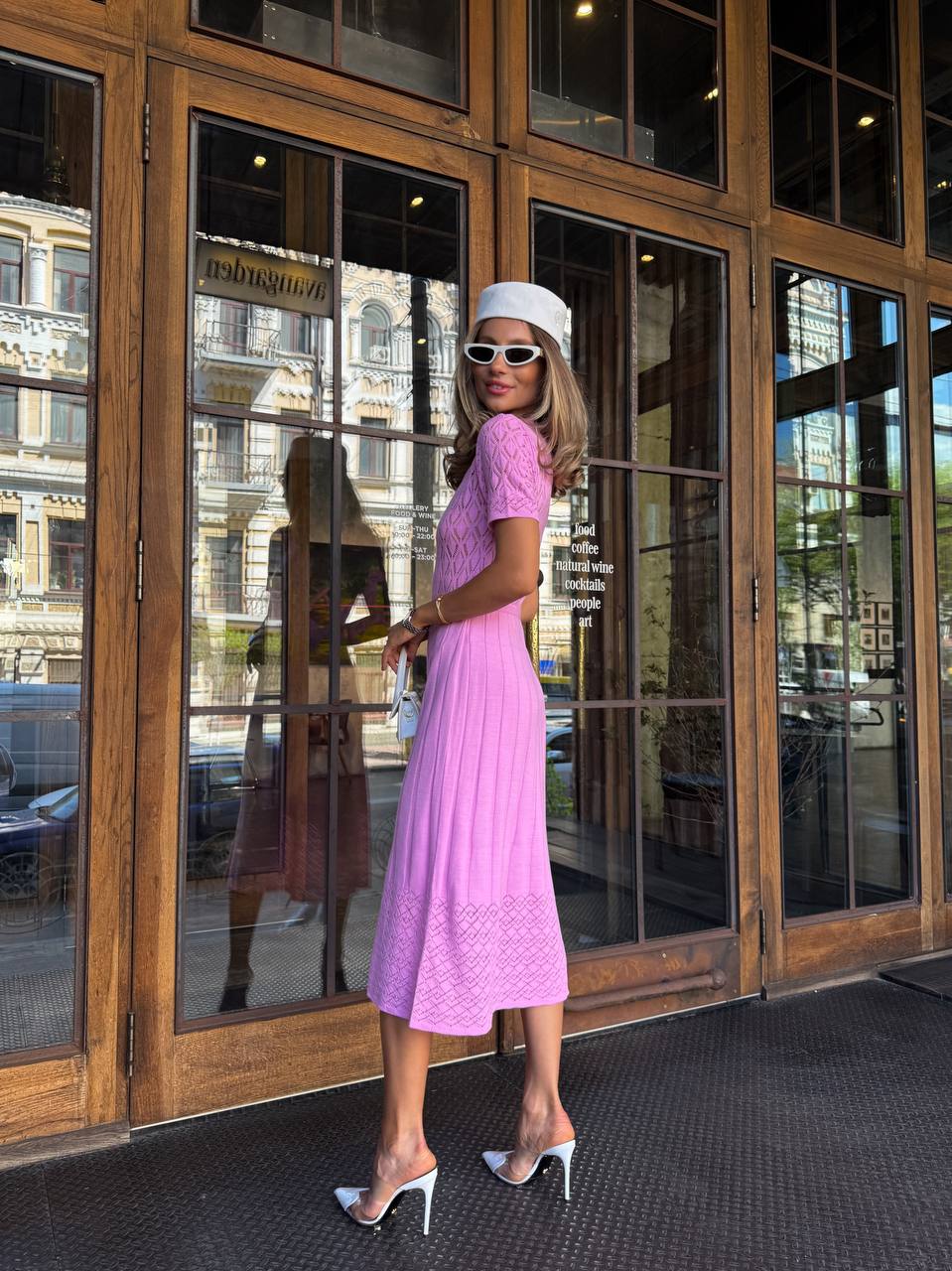 Woman in a pink dress and white hat standing in front of large glass doors.