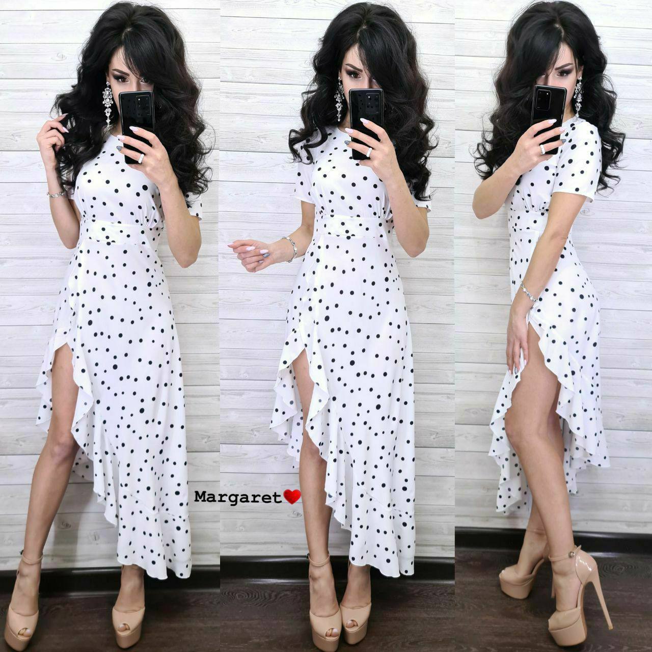 Woman wearing a white dress with black polka dots, posing in front of a white brick wall.