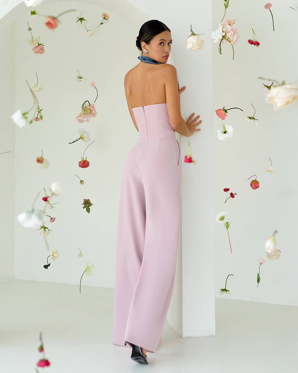 Woman in a pink strapless jumpsuit standing against a white wall with floral decorations.