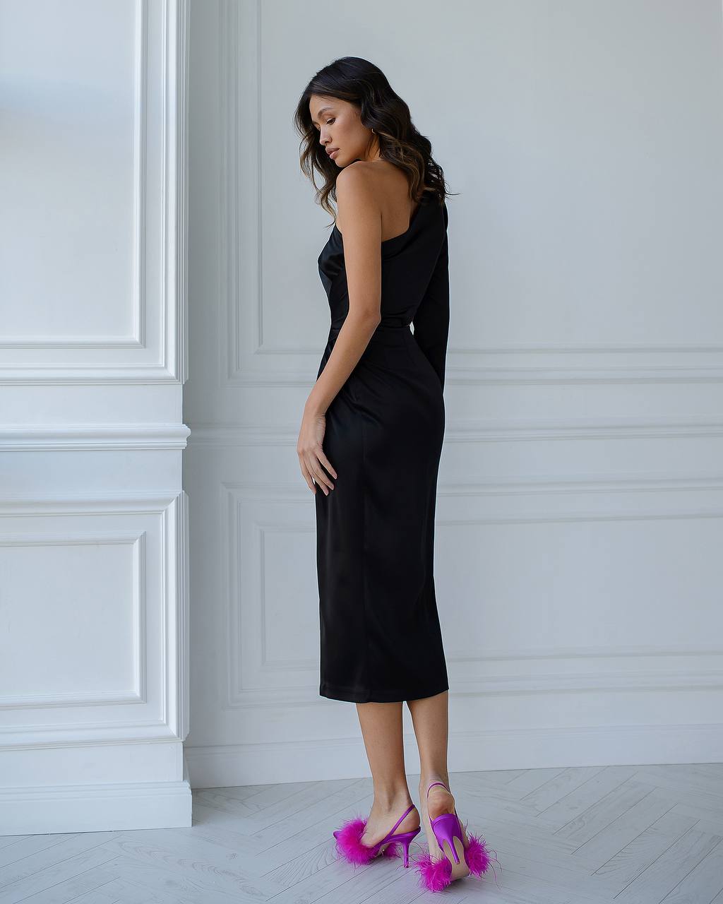 Woman in a black dress with pink sandals against a white paneled wall.