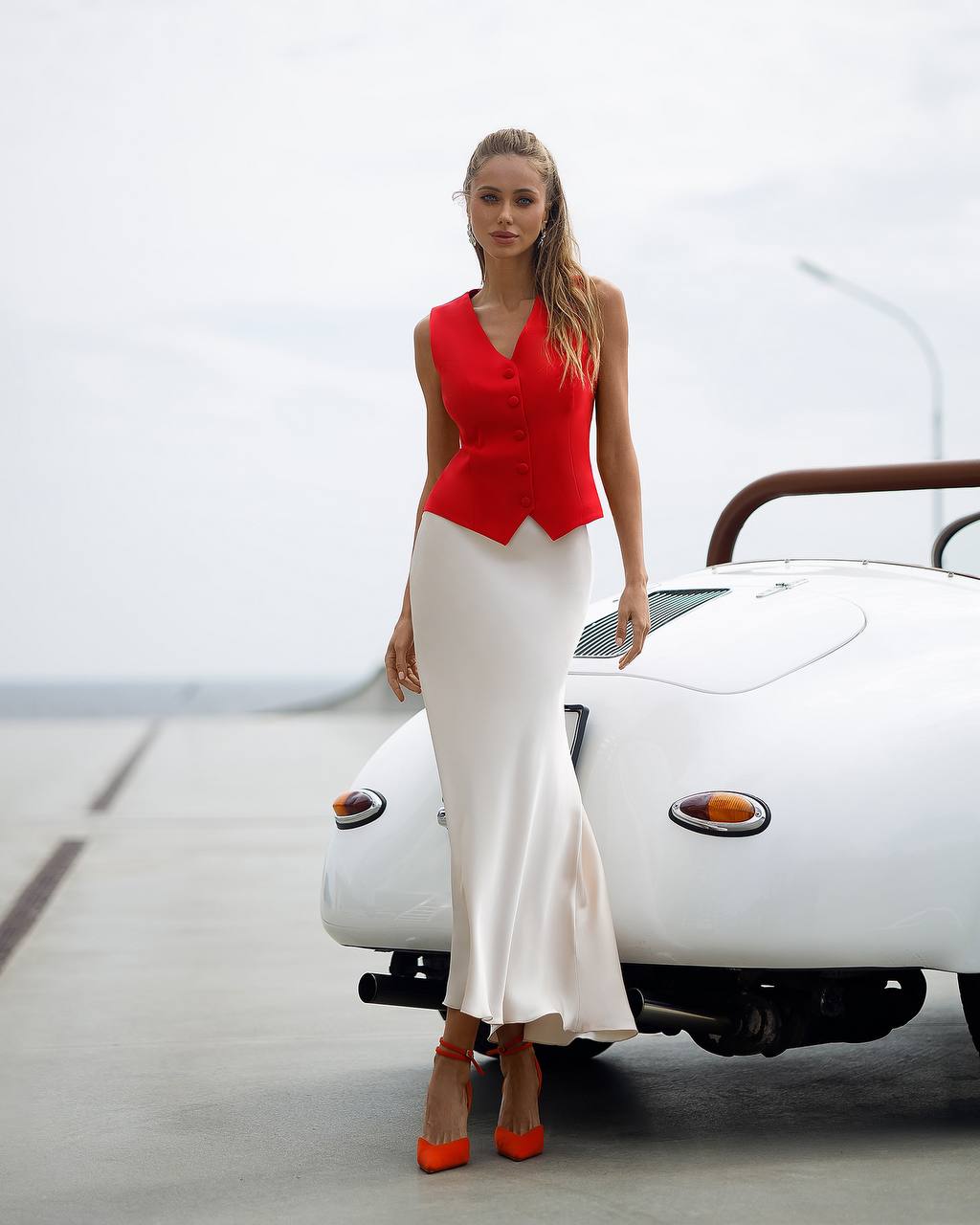 Woman in a red top and white skirt standing next to a vintage car.
