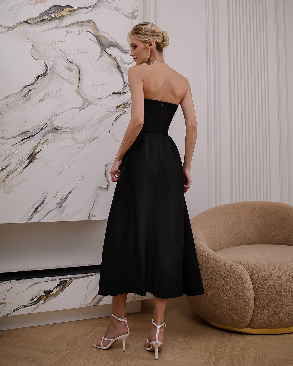 Woman in a black strapless dress standing in a room with marble wall and brown chair.