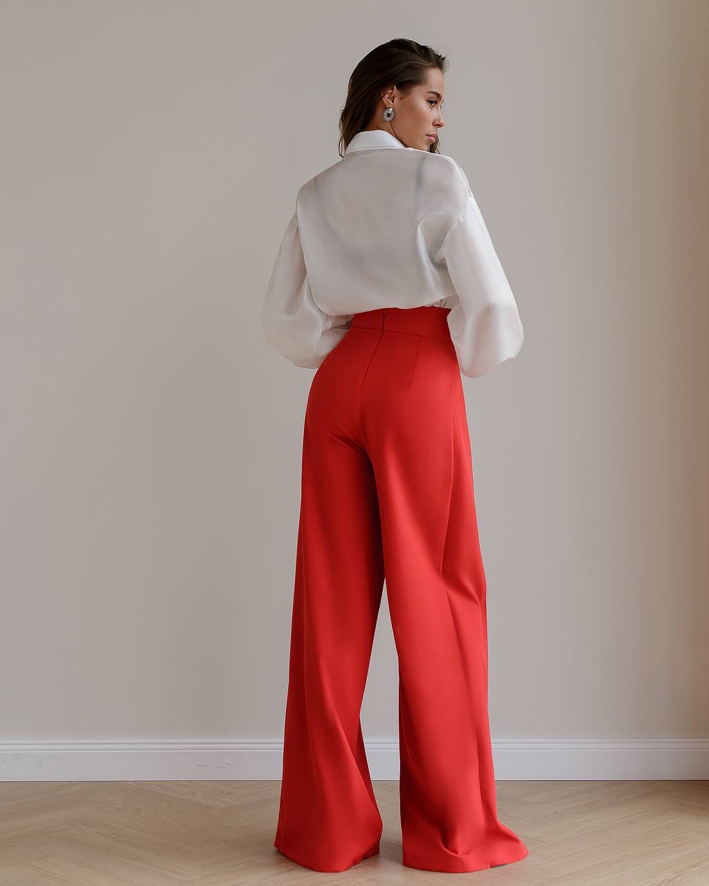 Person wearing a white blouse and red pants against a plain wall.