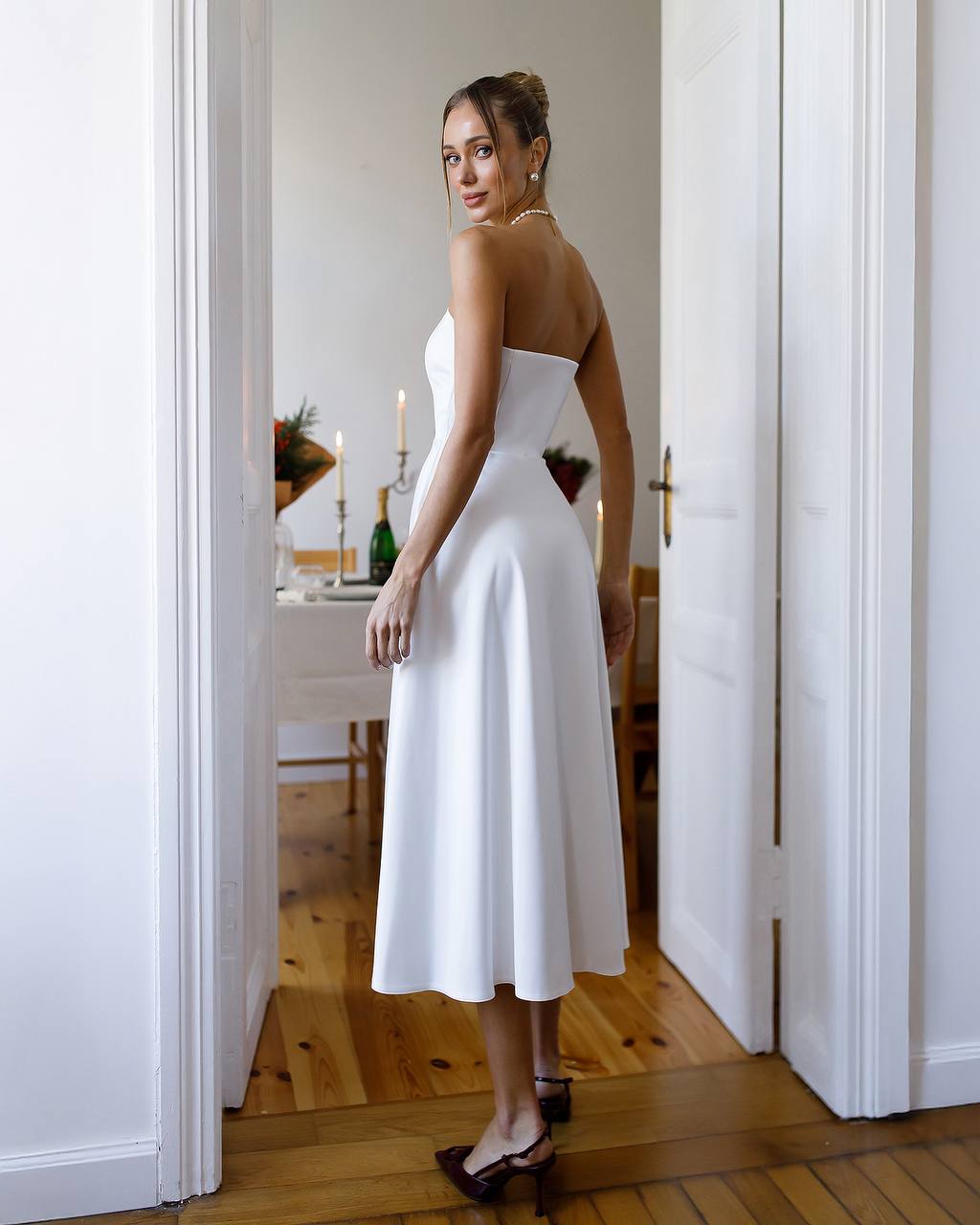 Woman in a white strapless dress standing in a room with wooden flooring and white walls.