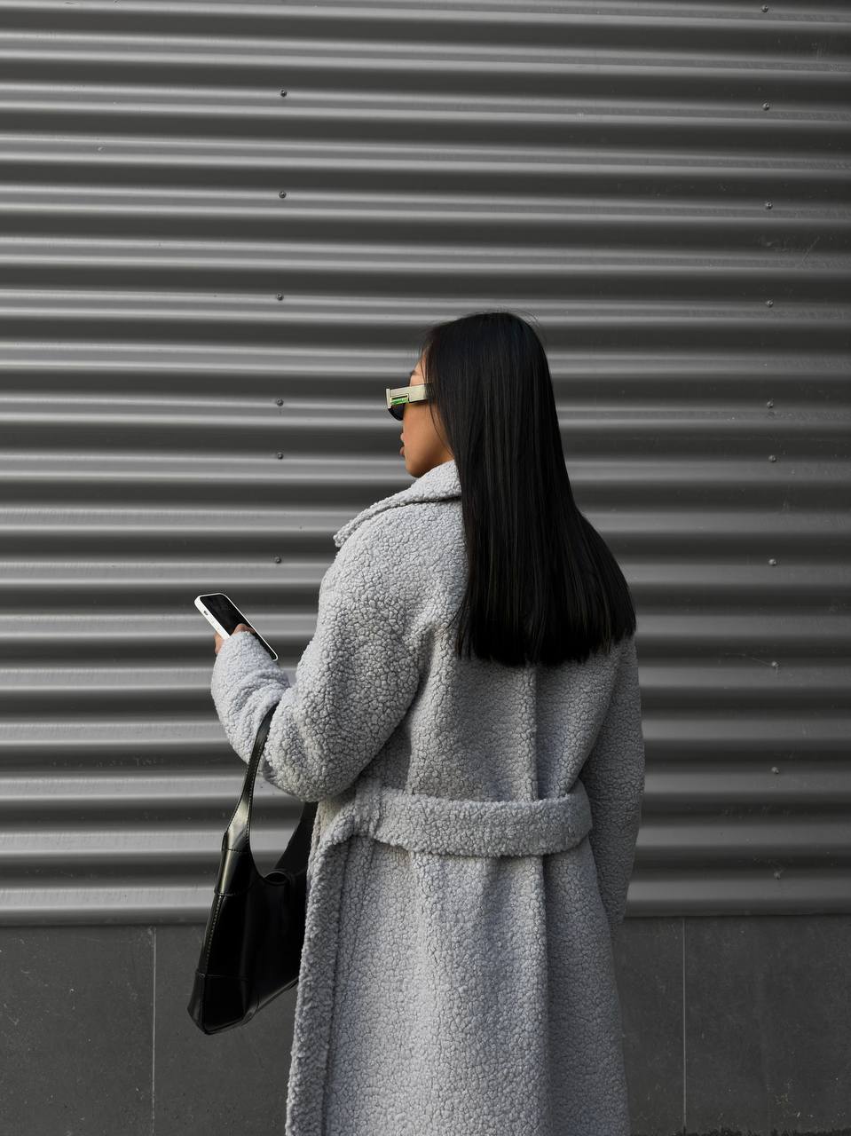 Person wearing a gray coat and holding a phone, standing in front of a metallic shutter.