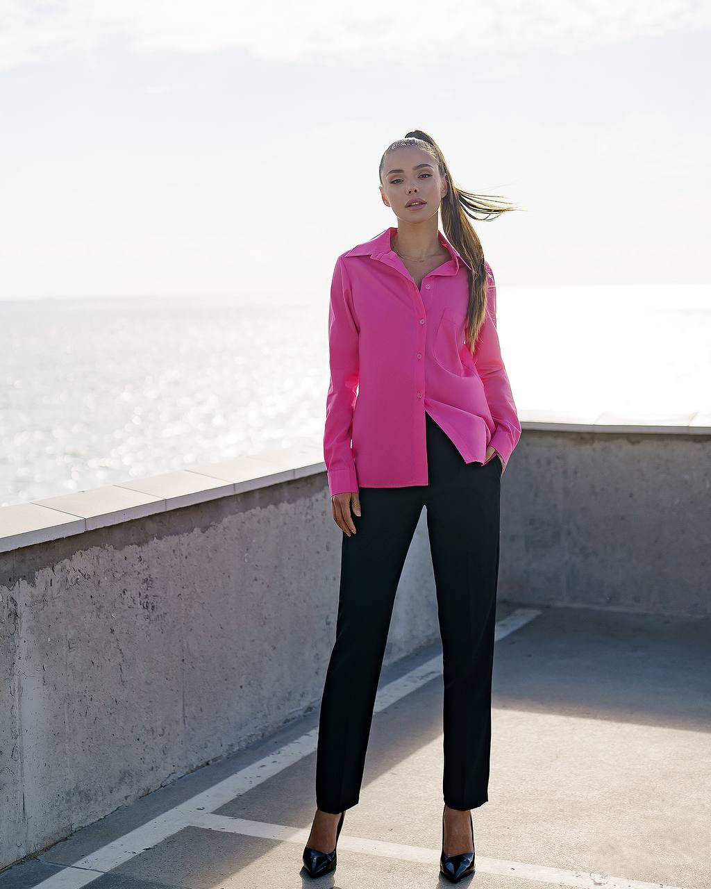 Woman wearing a bright pink shirt and black pants standing on a rooftop with a white wall in the background.