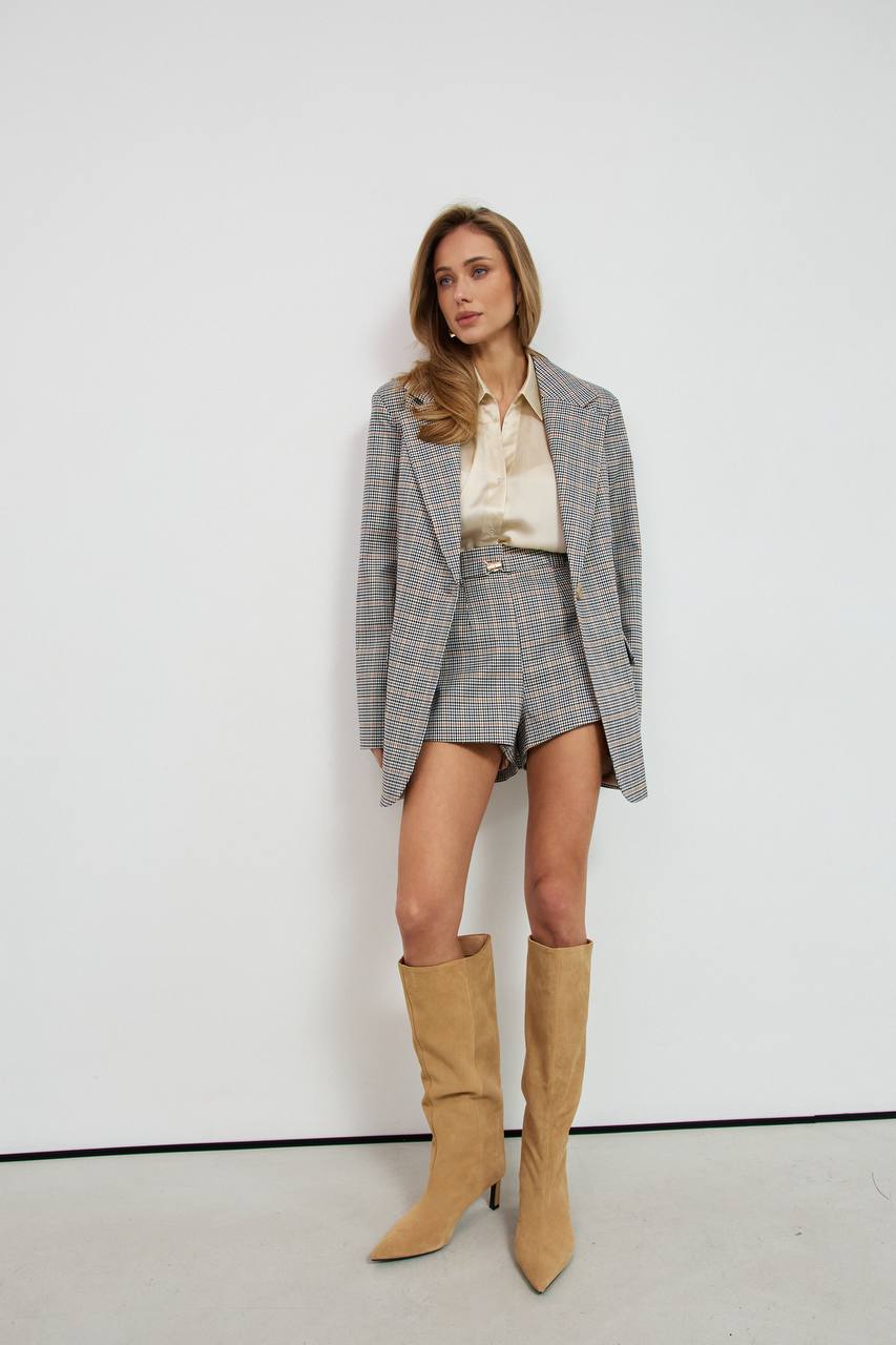 Woman wearing a gray checkered blazer, beige shirt, shorts, and tan knee-high boots against a white background.
