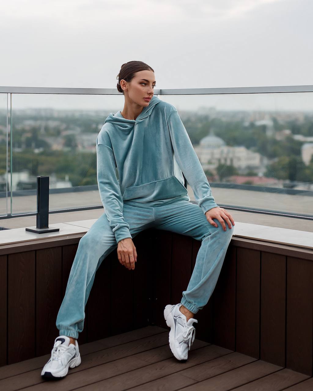 Person wearing a light blue tracksuit sitting on a ledge with a cityscape view.