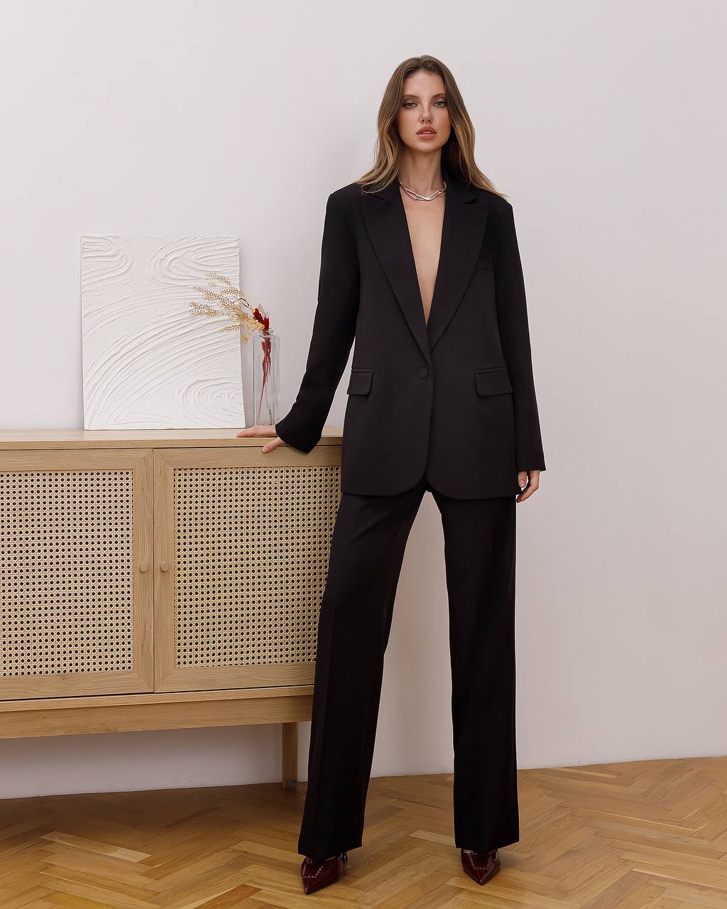 Woman wearing a black suit standing in a room with wooden flooring and a light-colored wall.