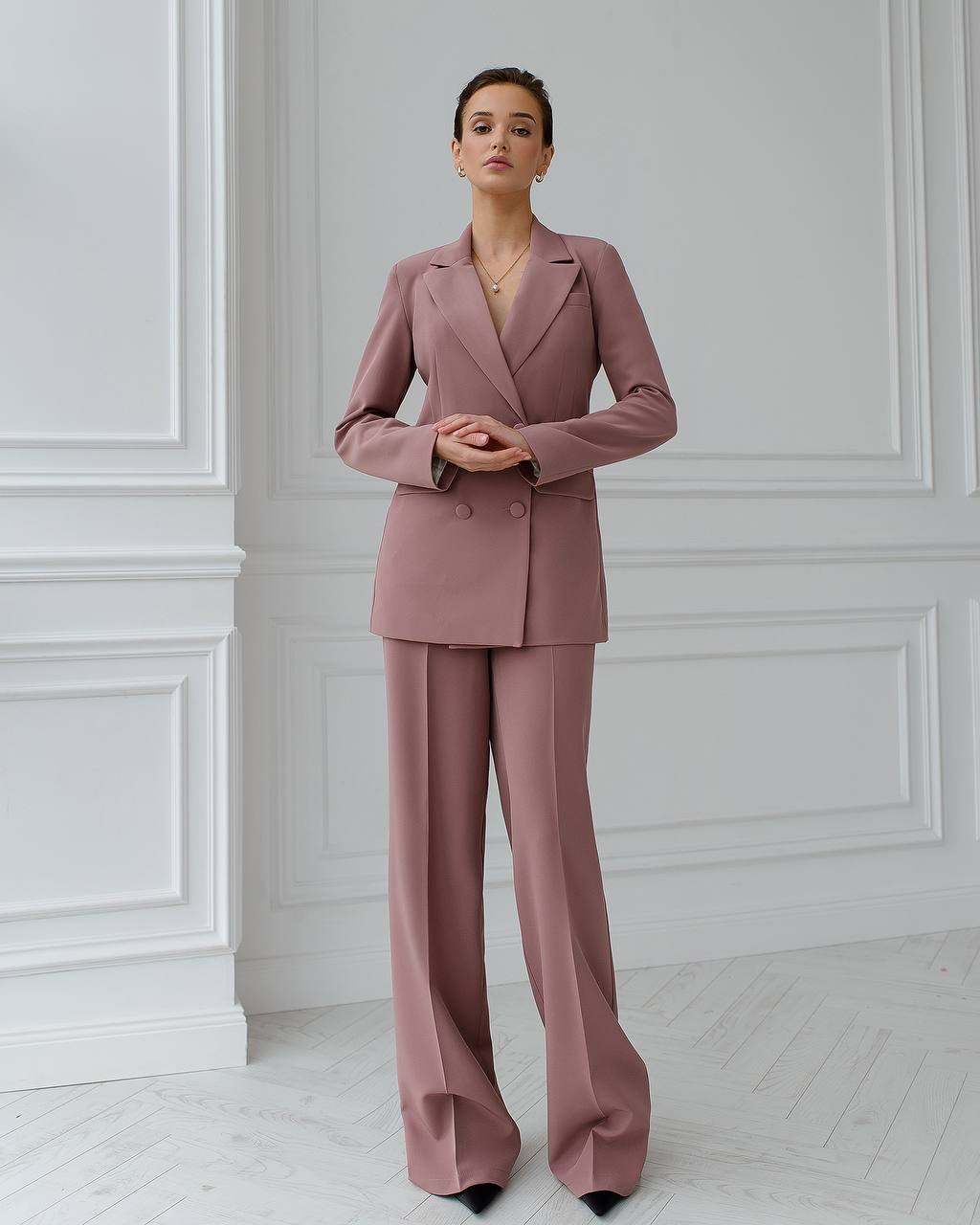 Woman wearing a pink suit standing against a white paneled wall.