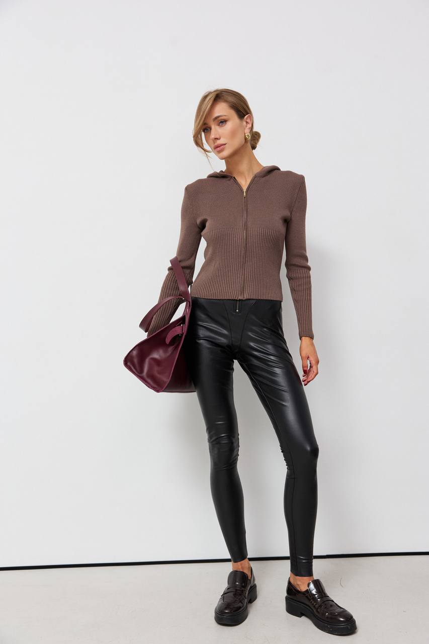 Woman wearing a brown zip-up hoodie, black leather pants, and holding a purple handbag against a white background