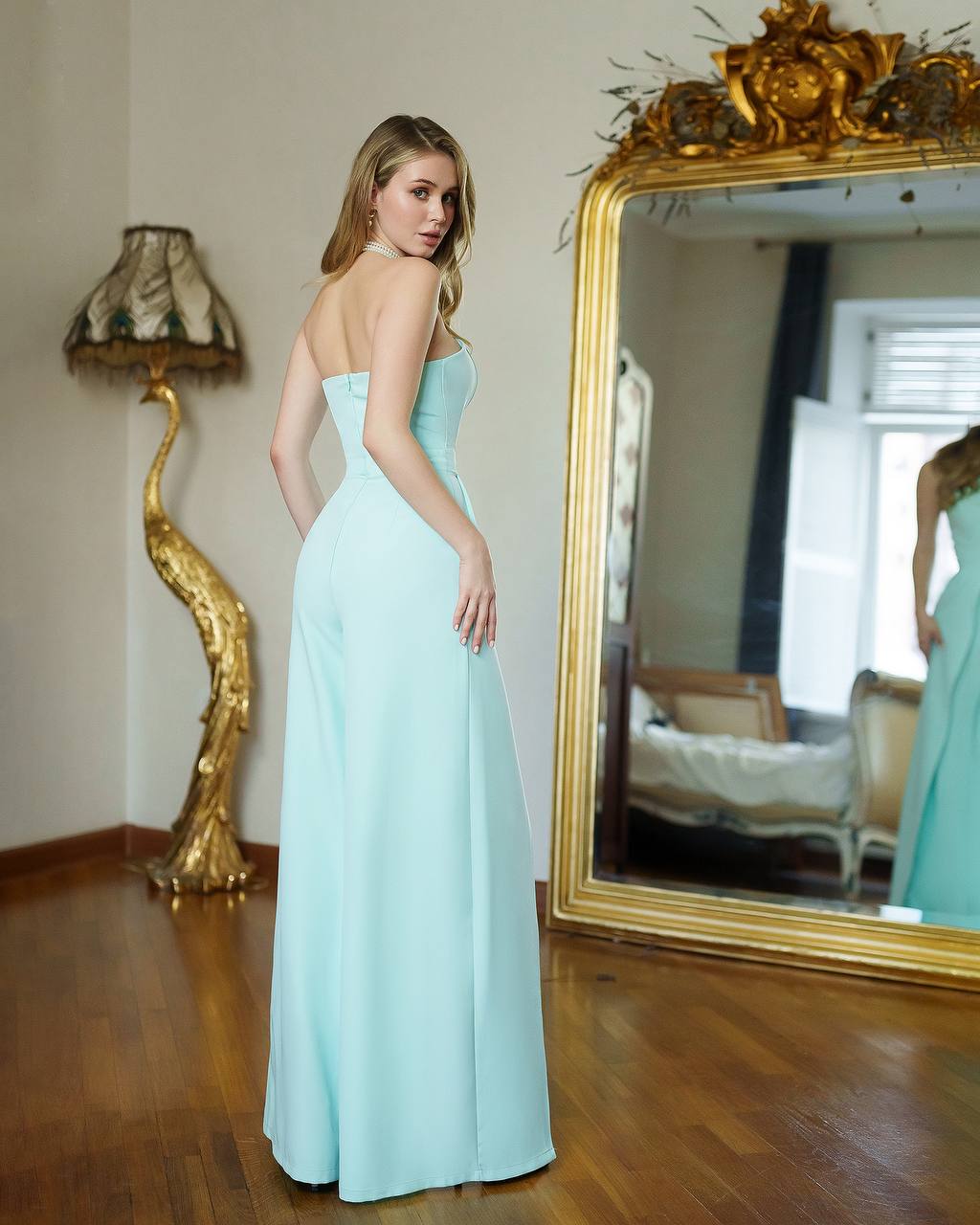 Woman in a light blue dress standing in a room with a large mirror and decorative lamp.