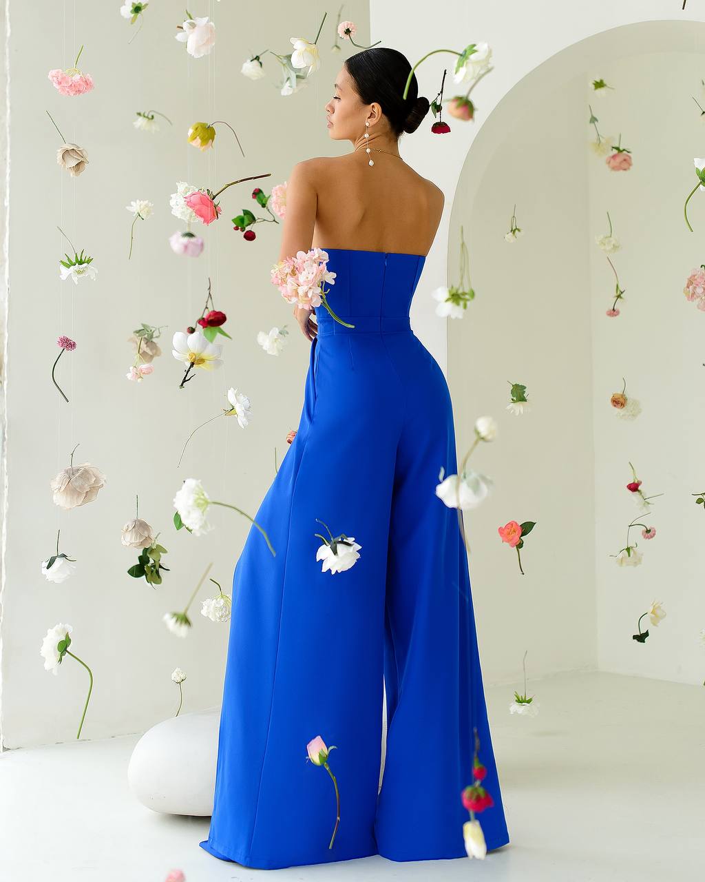 Woman in a blue strapless jumpsuit with floral decorations against a floral wall.