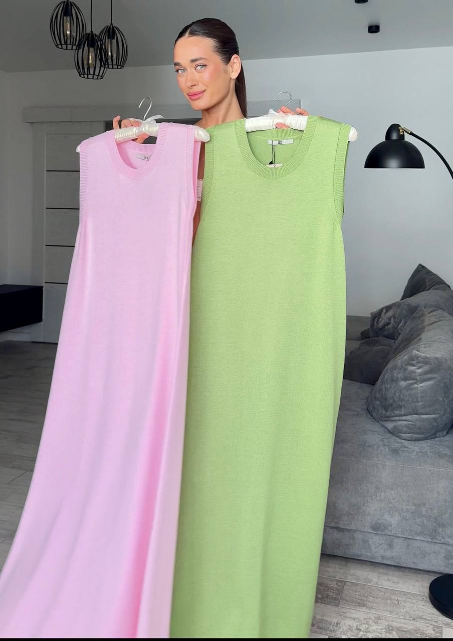 Woman holding two dresses, one pink and one green, in a room with a gray sofa and lamp.