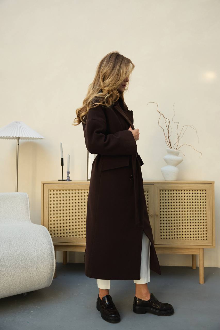 Woman wearing a long dark coat in a room with a lamp and cabinet.