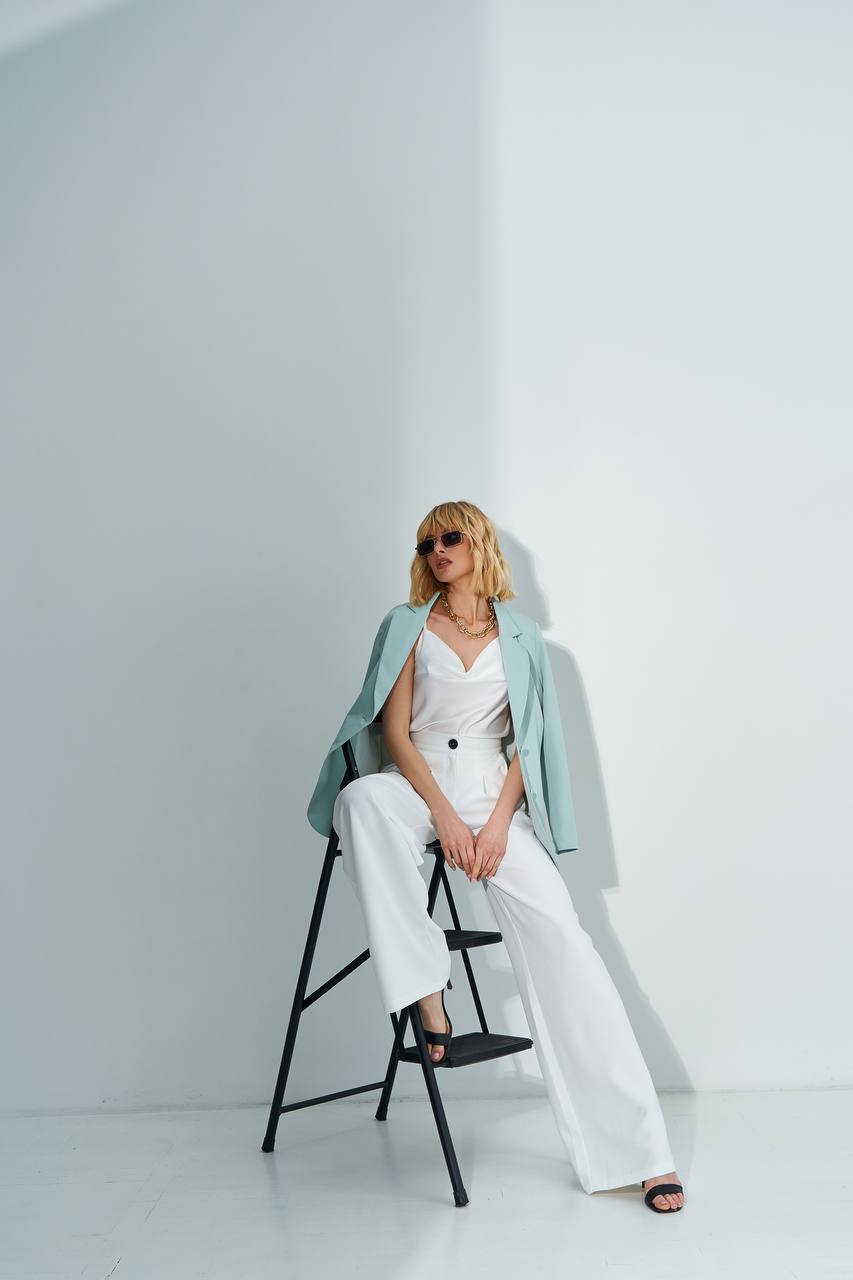 Woman in a white outfit with a light blue jacket sitting on a black stool against a white background
