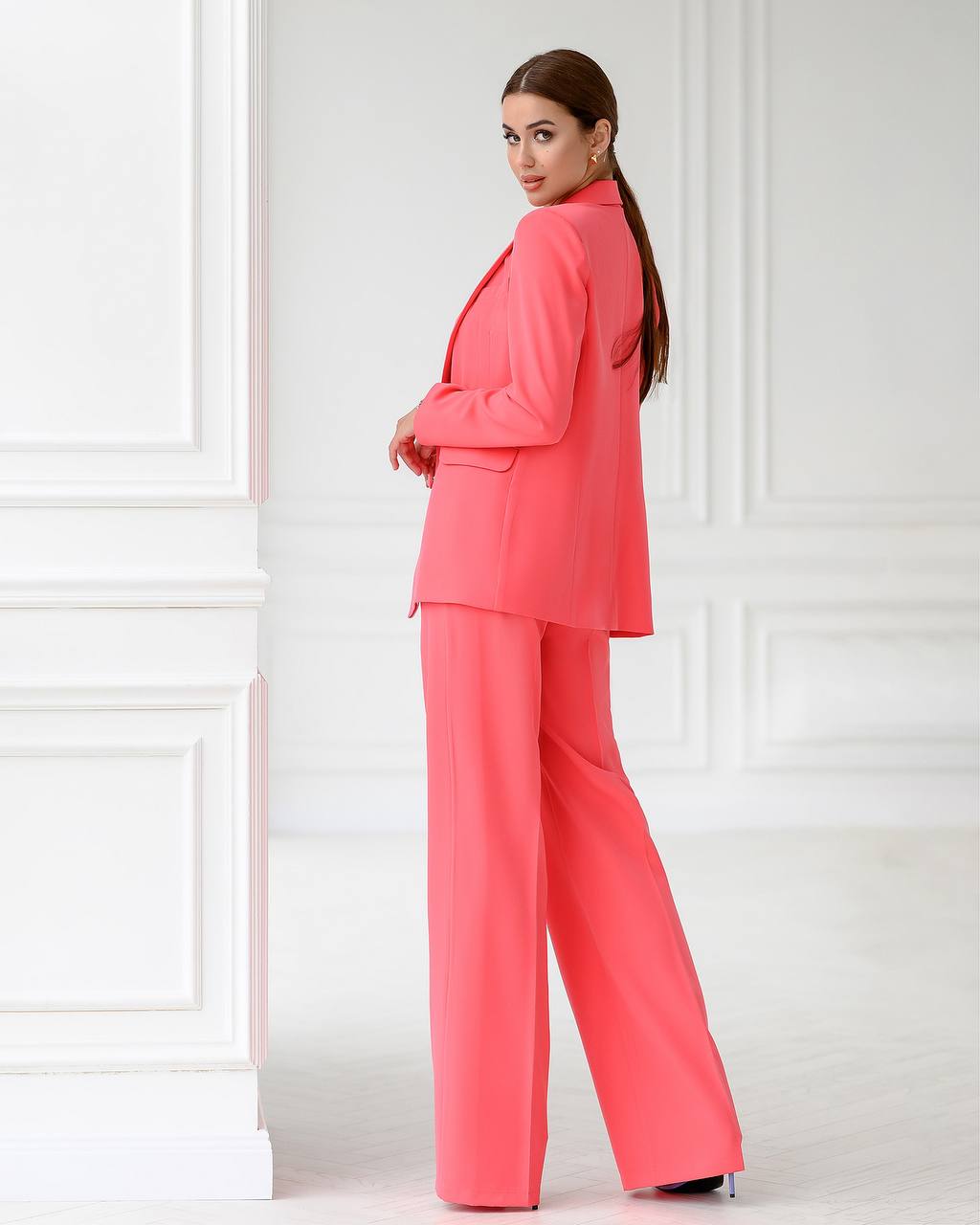 Woman wearing a coral suit standing against a white paneled wall.