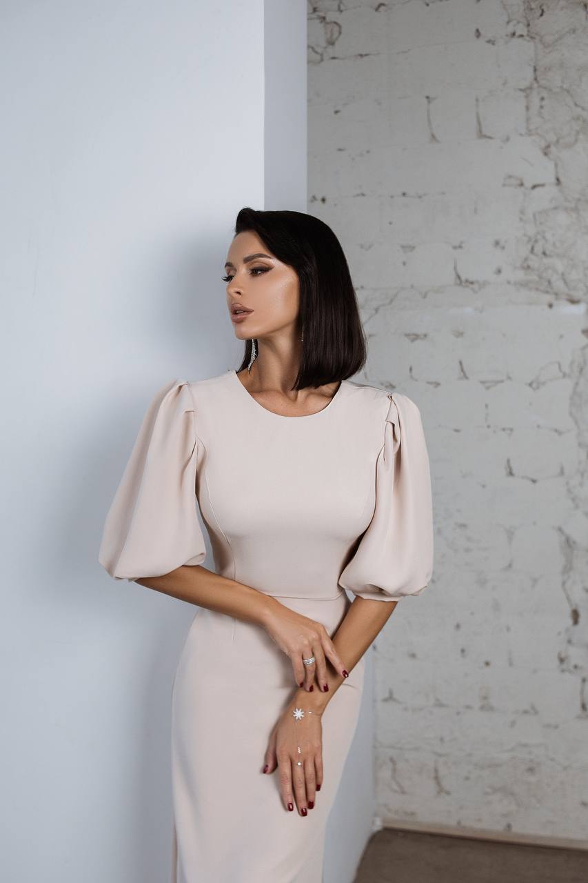 Woman wearing a beige dress with puffed sleeves against a light gray wall.