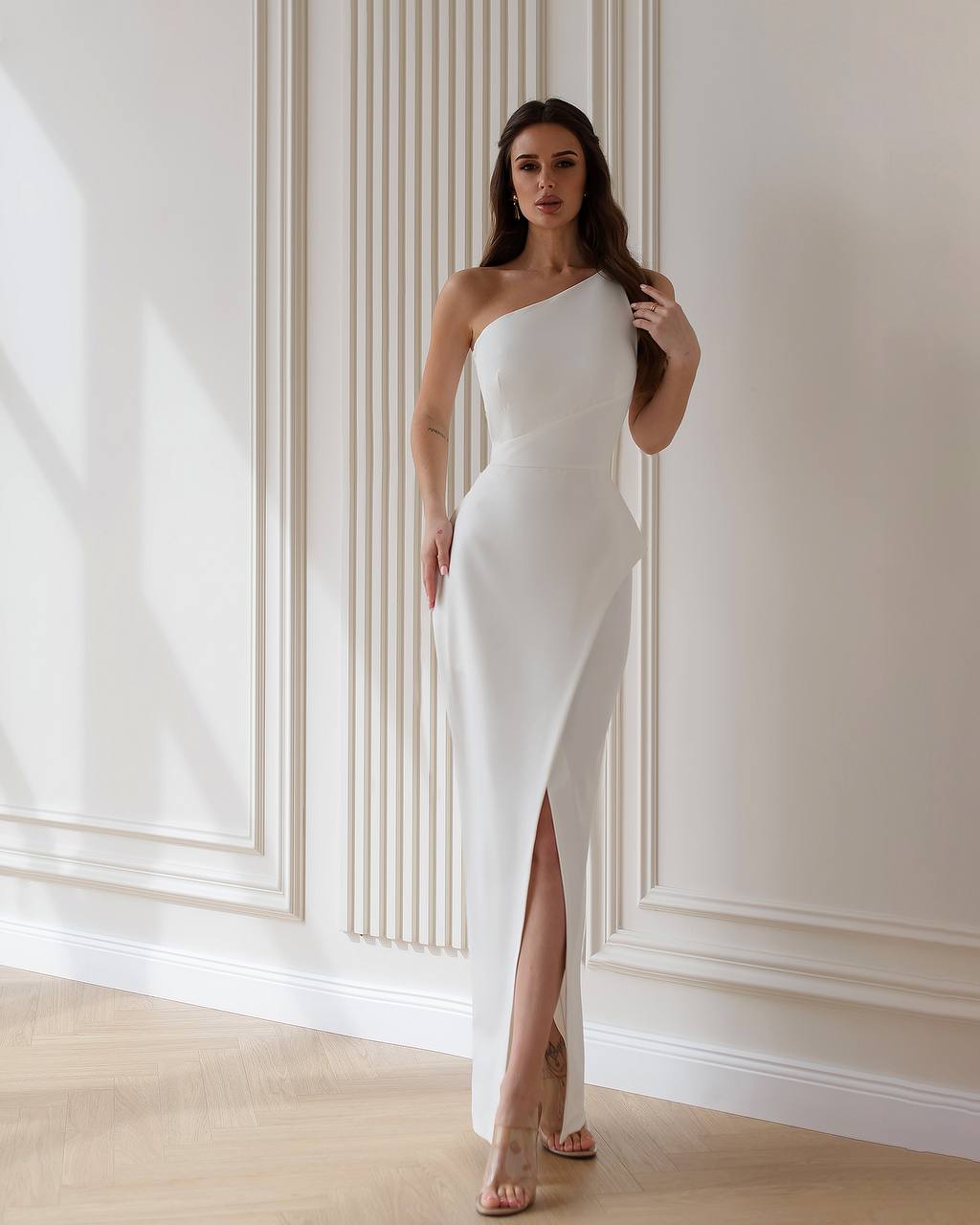 Woman wearing a white one-shoulder dress with a high slit in a room with white walls and wooden floor.