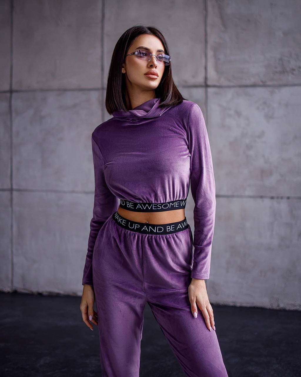 Woman wearing a purple outfit with text on the waistband against a gray tiled wall.