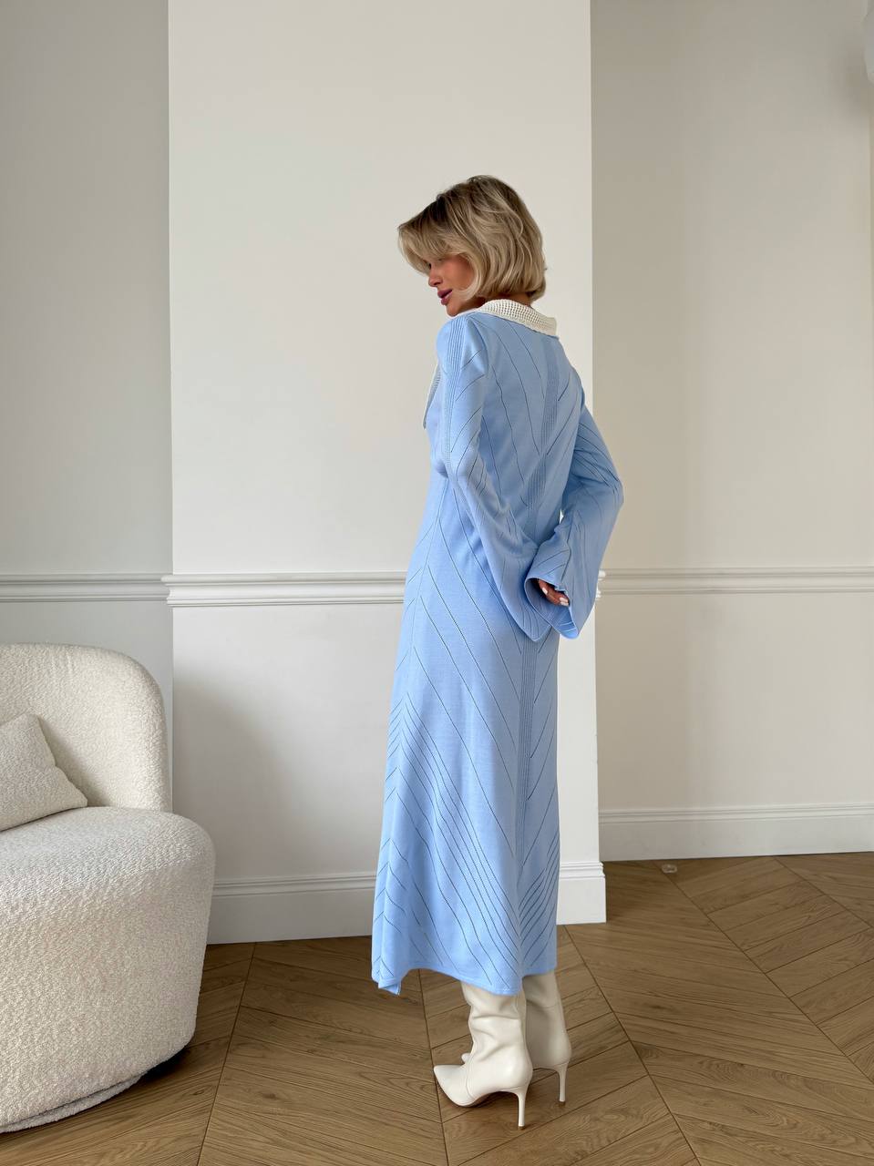 Woman wearing a light blue dress standing in a room with a white wall and wooden floor.