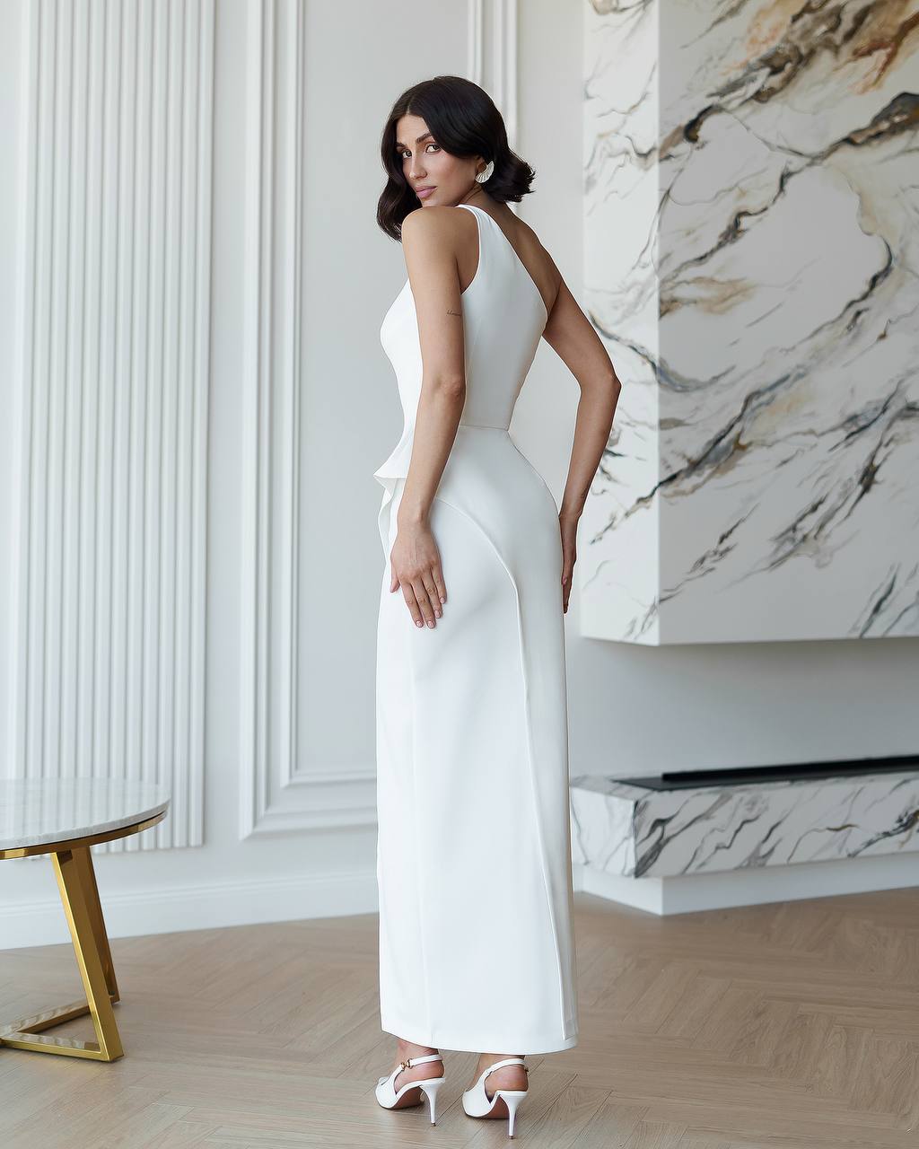 Woman in a white dress standing in a stylish interior setting with marble wall.